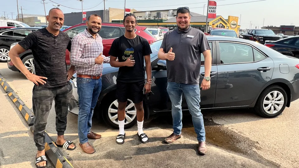 Four people beside a silver sedan in the vehicle lot